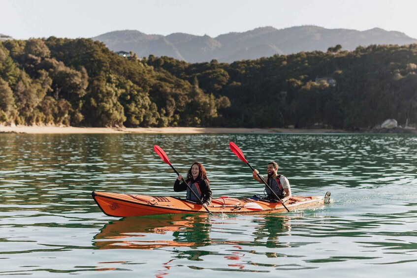 Two people kayaking on a calm lake with a forested shoreline and mountains in the background. 2