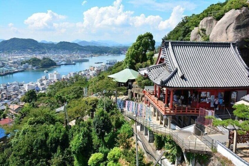 A traditional Japanese building with a tiled roof, surrounded by lush greenery and overlooking a river with a cityscape in the background. 1