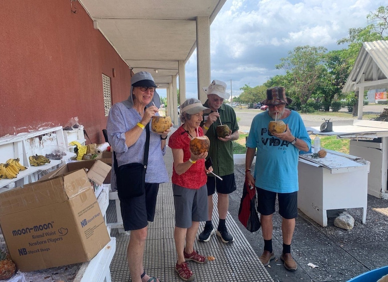 A group of people outdoors, some holding coconuts, with a table of bananas and a cardboard box labeled 'moon-moon Round Waist Newborn Diapers' in the background. 3