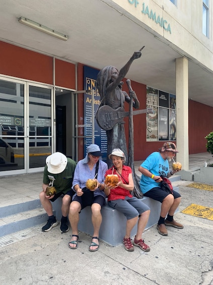Four people sitting on a bench outside a building with a statue of a musician. 4