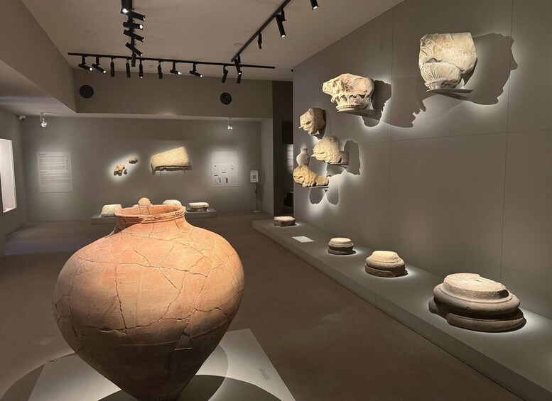A museum interior with pottery and stone artifacts displayed on walls and a large vase in the foreground. 1