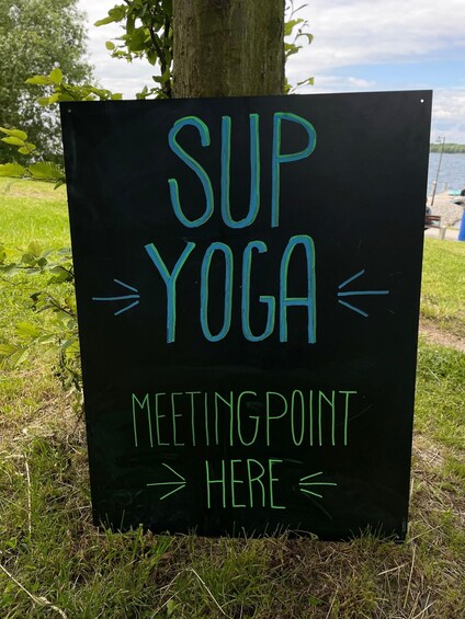 A sign with 'SUP YOGA MEETINGPOINT HERE' written on it, placed on grass near a tree. 1