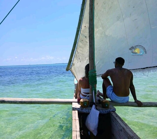 A sailboat with two people on board, one sitting and one standing, with clear turquoise water and a blue sky in the background. 1