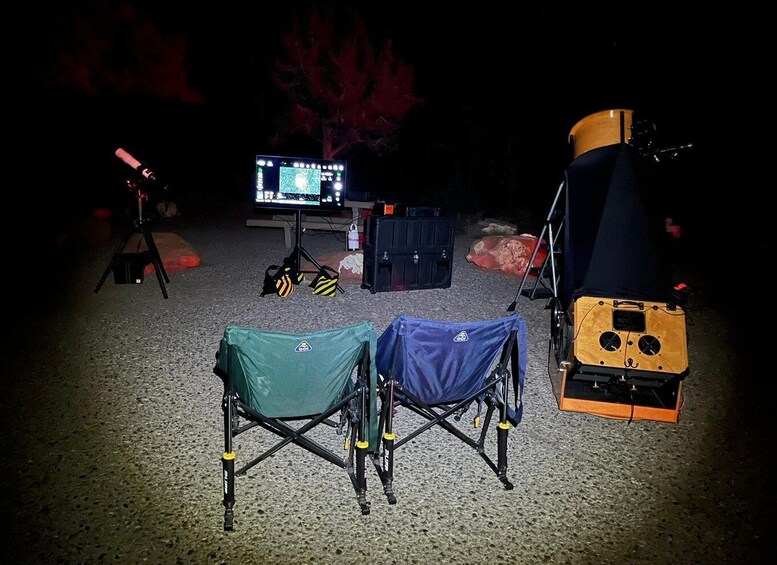 A camping setup with a telescope, a computer monitor, and two portable chairs. 2