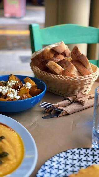 A table with a blue bowl of roasted potatoes and feta cheese, a wicker basket of bread, and a plate with a yellow dish. 2