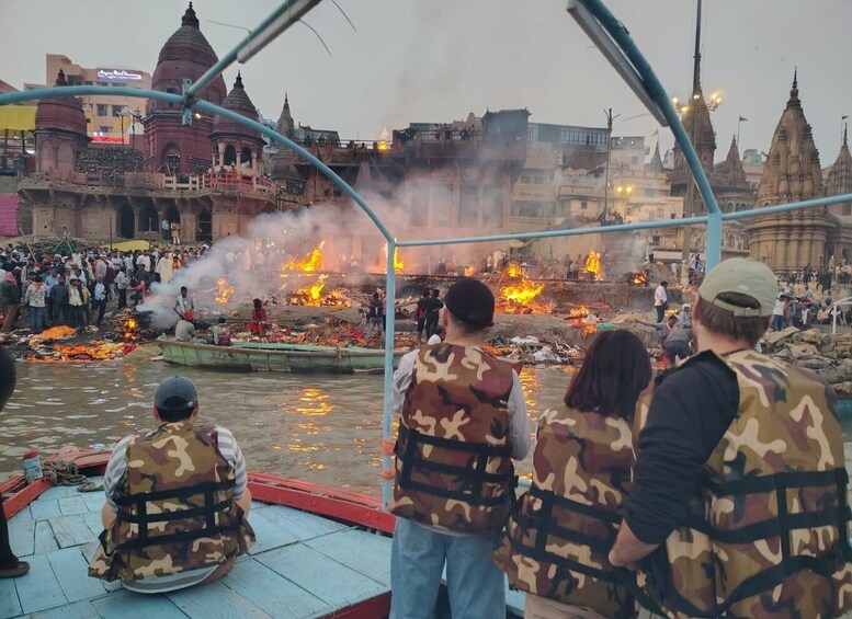 A riverbank with fires and smoke, surrounded by people and traditional architecture. 1