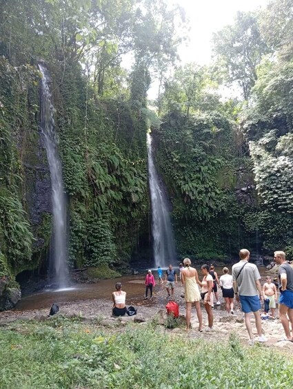 A waterfall surrounded by lush greenery with people walking and sitting around. 1