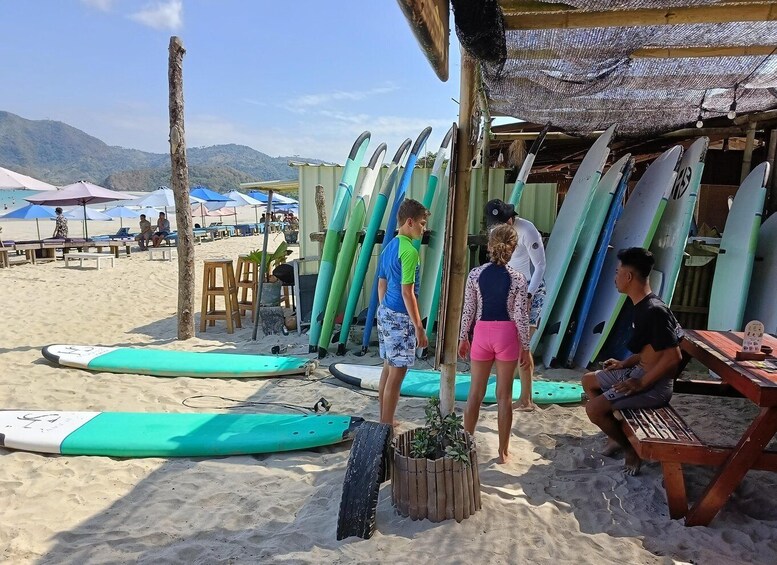 A beach with surfboards lined up under a shelter, people standing and sitting, and a mountainous landscape in the background. 5