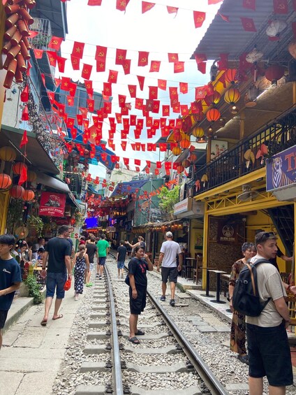 A street with red flags and lanterns, a railway track, and people walking. 1