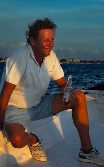 A person sitting on a boat, holding a bottle, with water and buildings in the background. 5