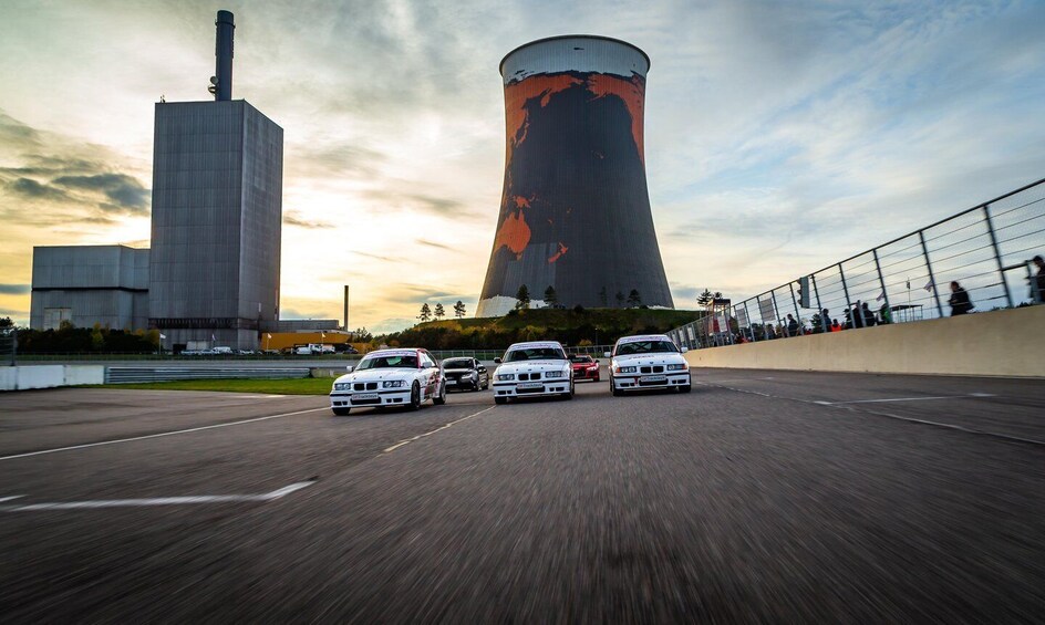 A racing track with four white cars, a cooling tower, and a tall building in the background. 2