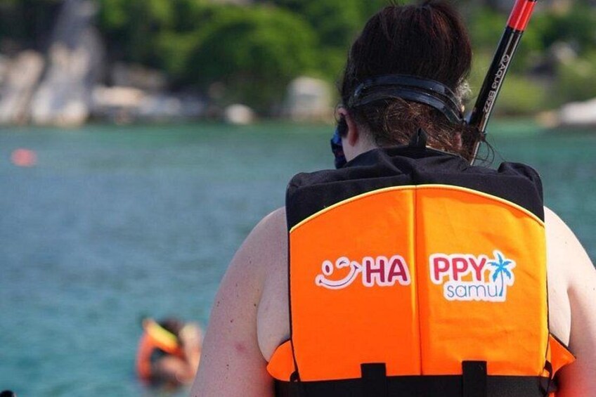 A person wearing an orange life jacket with HA and Ppy samui logos, holding a red and black paddle, in a body of water with a blurred background. 5