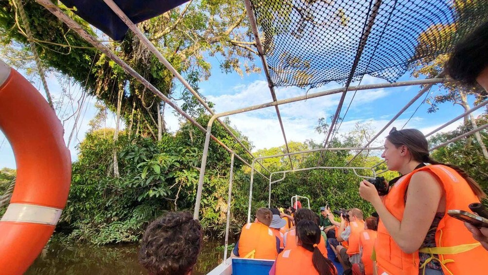 A group of people in orange life vests on a boat tour through a forested area with a canopy overhead. 3