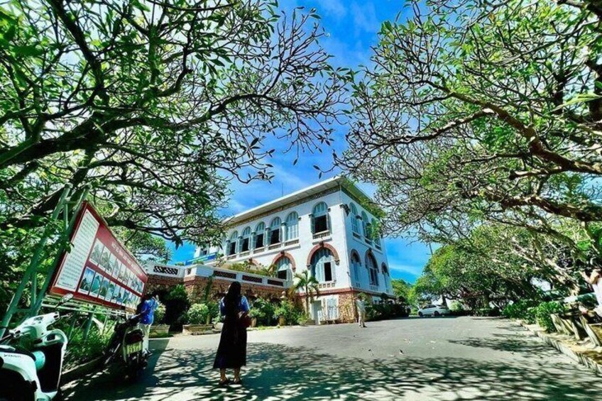 A historic building with arched windows and a balcony, surrounded by trees and a signboard. 1
