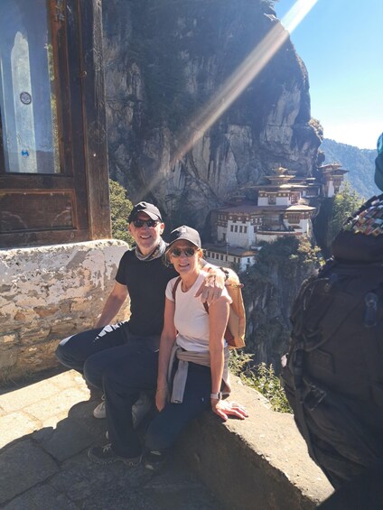 Two people sitting on a ledge with a mountainous landscape and traditional buildings in the background. 2