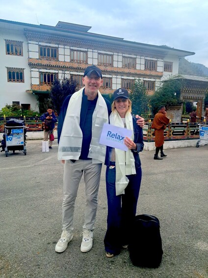 Two people standing in front of a building with traditional architecture, holding a sign that says 'Relax Getaways.' 1