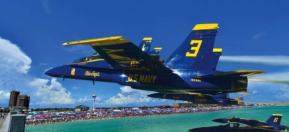 A US Navy Blue Angels aircraft flying over a crowded beach with a clear blue sky. 1