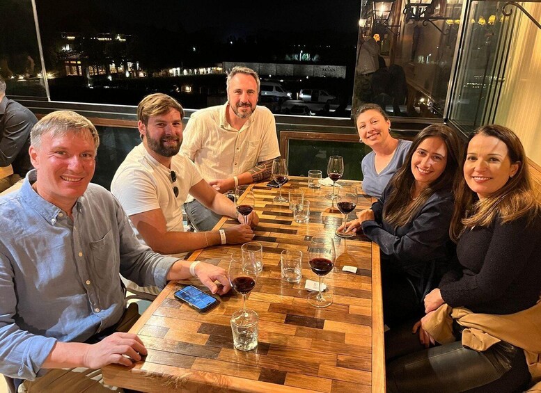 A group of people seated at a wooden table with wine glasses, in a room with a view of a cityscape at night. 5