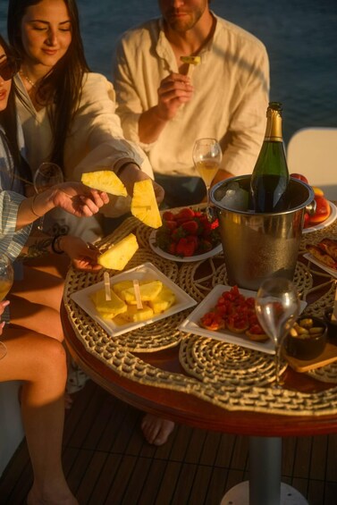 A group of people enjoying a meal on a boat with tropical fruits, drinks, and a bottle of champagne. 5