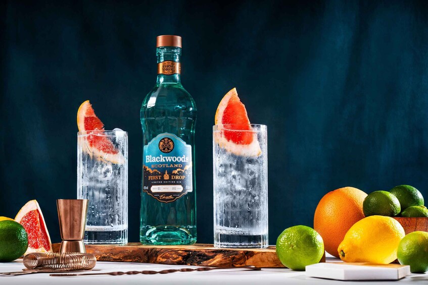 A bottle of Blackwoods Scotland First Drop gin, two glasses with ice and citrus slices, and fresh limes and oranges on a wooden board. 1