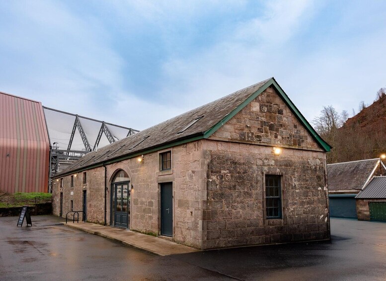 A stone building with a green roof and arched windows, located next to a metal structure and a hill in the background. 4
