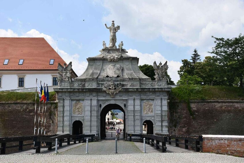 A historic stone archway with statues and flags, leading to a building with a red roof. 3