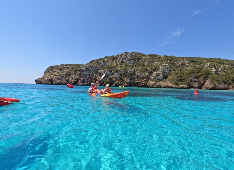 Two people kayaking on clear blue water with a rocky island in the background. 1