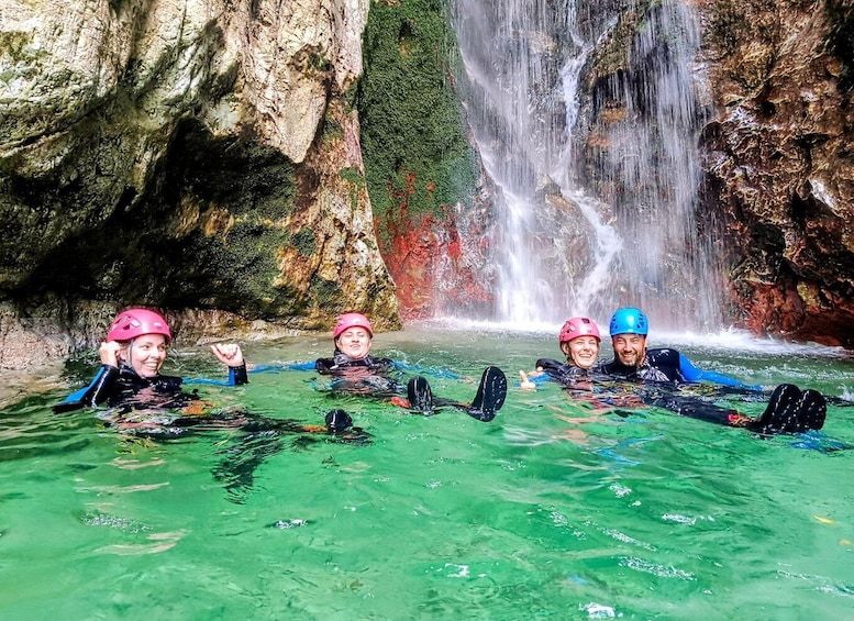 Four people in water with kayaks, a waterfall, and rocky cliffs. 1