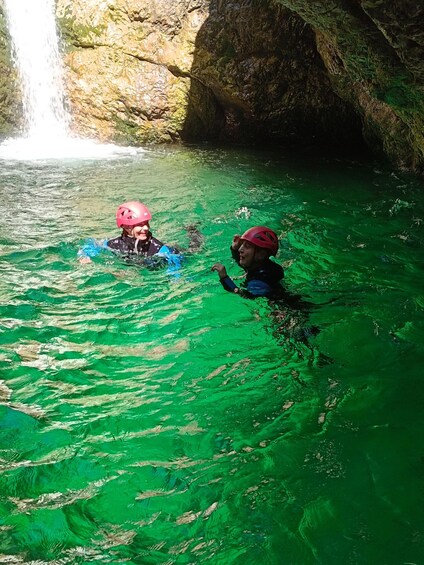 Two people in a clear, green body of water with a waterfall in the background. 2