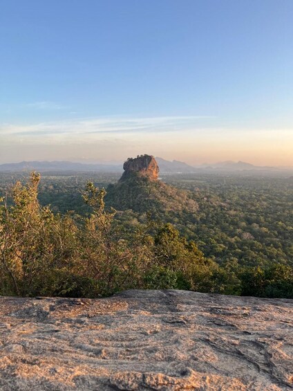 A rocky outcrop with a prominent rock formation on a hill, surrounded by dense forest and a distant cityscape under a clear sky. 5
