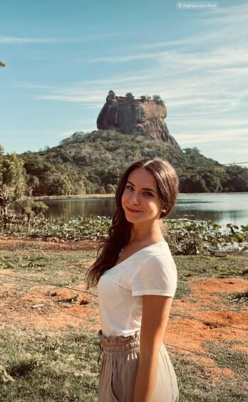 A woman standing in a natural landscape with a lake and a prominent rock formation in the background. 1