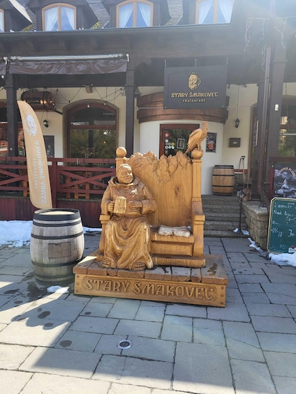 A wooden statue of a man holding a book, a wooden chair, and a sign that reads 'STARY ŠMAKOVEC restaurant' in front of a building with windows and a wooden fence. 2