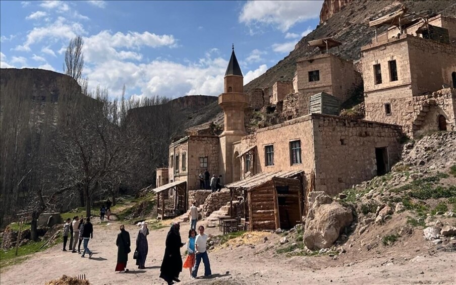 A historic stone building with a tower, surrounded by a mountainous landscape and a dirt path with people walking. 5