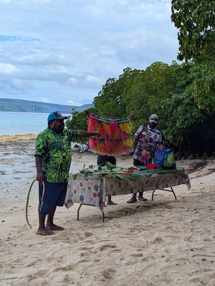 Two people on a sandy beach with a table covered in a patterned cloth, holding colorful woven items. 5