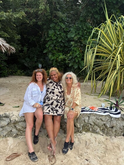 Three people sitting on a stone wall by a sandy beach with tropical vegetation in the background. 2