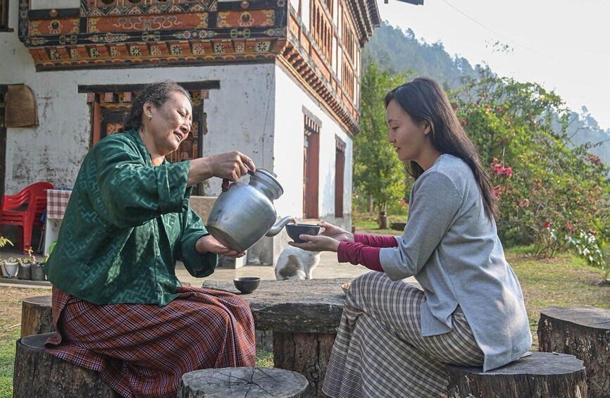 Two people sitting on wooden stumps outdoors, one pouring tea from a metal pot into a cup. 3