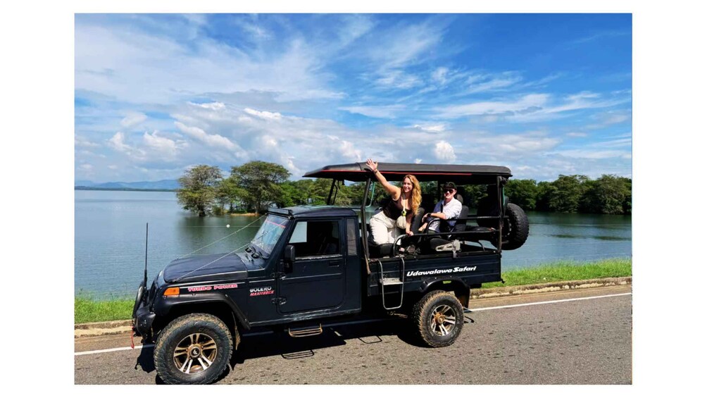A black Udawala Safari vehicle with two passengers on a road beside a lake. 5