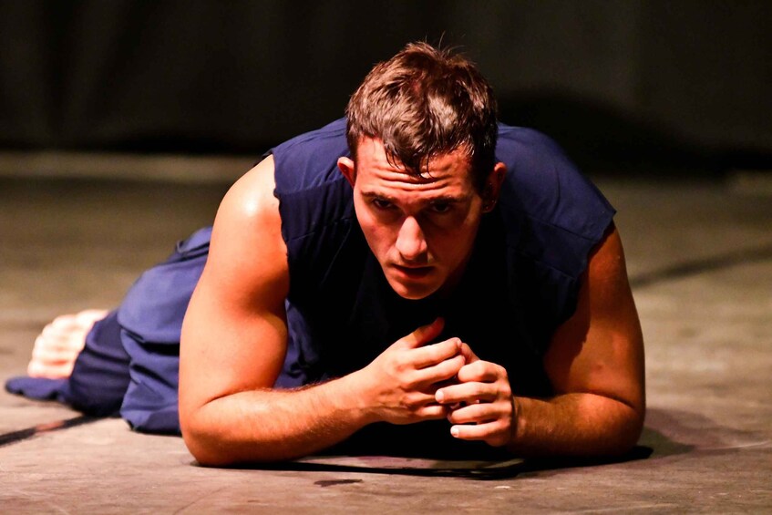 A person lying prone on a stage with a dark background. 4
