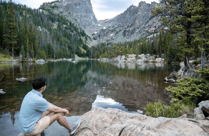 A man sitting by a clear mountain lake surrounded by dense forest and rocky terrain. 3