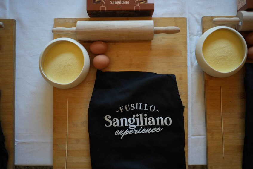 A kitchen counter with a rolling pin, two bowls of flour, eggs, and a black cloth with 'FUSILLO Sangiliano experience' printed on it. 5