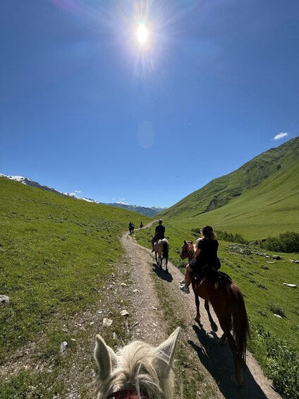 A group of people horseback riding on a dirt trail through a green valley with mountains in the background. 4