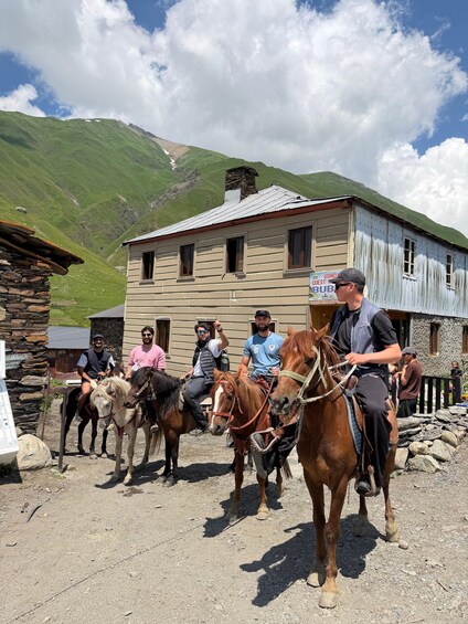 A group of people riding horses in a rural area with a stone building and a mountain in the background. 2