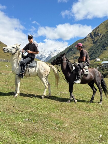 Two people riding horses in a grassy field with mountains in the background. 1