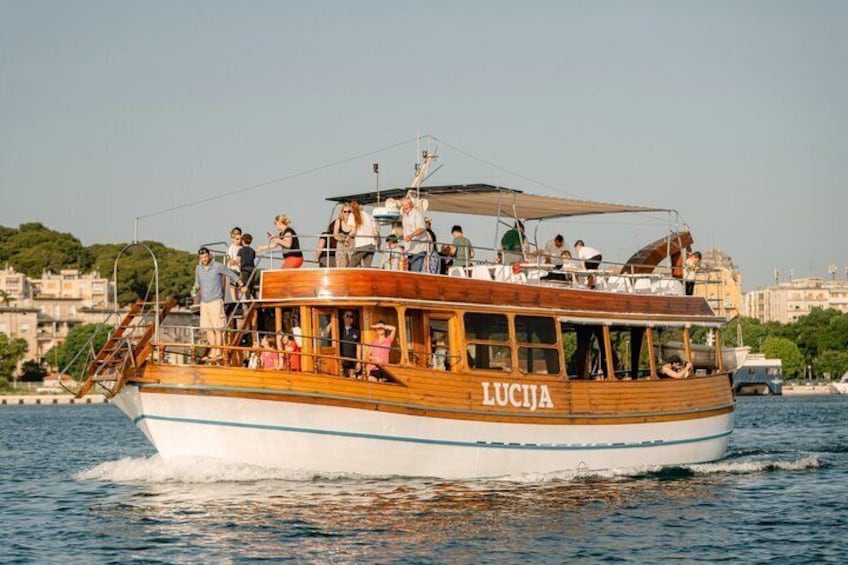 A wooden boat named 'LUCJA' with passengers on board, sailing on a body of water with a cityscape in the background. 3