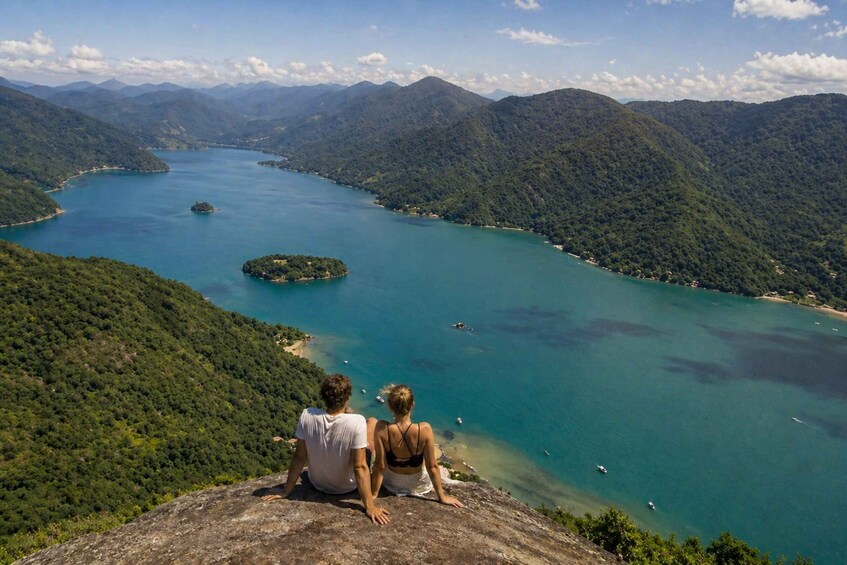 A couple sitting on a cliff overlooking a turquoise lake surrounded by forested mountains. 3