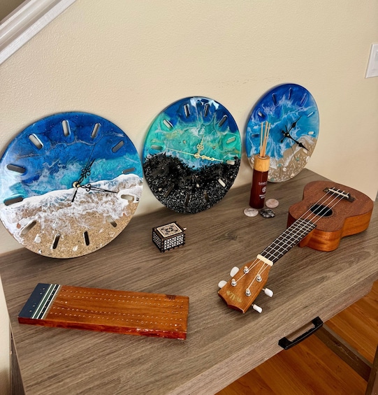 Three decorative clocks with ocean-themed designs, a small wooden box, a ukulele, and a wooden tray on a wooden table. 1