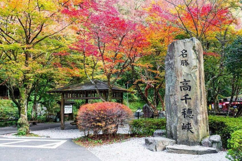 A stone monument with Japanese characters, surrounded by autumn-colored trees and a traditional Japanese structure. 2