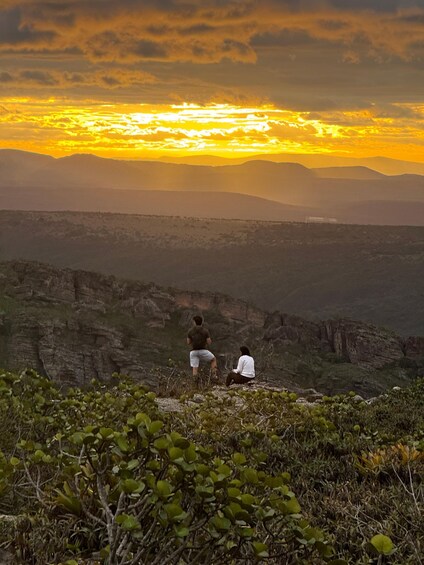 Two people sitting on a hill overlooking a valley with a sunset in the background. 2