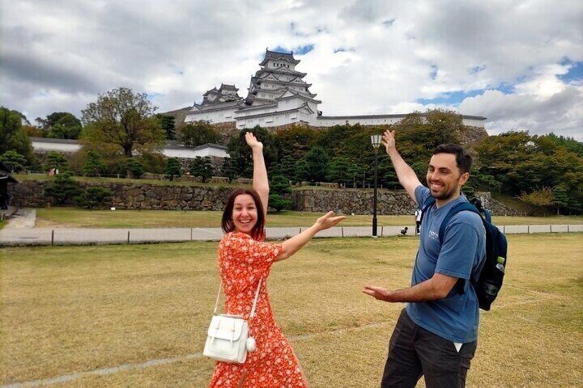 A person in a blue shirt and another in a red dress with a white bag, standing in a grassy area with a traditional Japanese castle in the background. 5