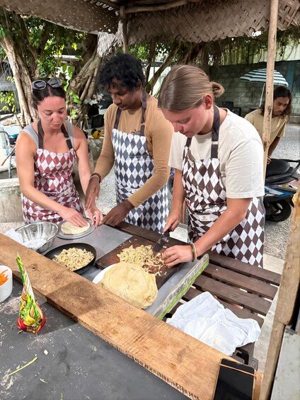 Three people preparing food outdoors, wearing aprons, with a table, a cutting board, and a bowl of shredded ingredients. 1
